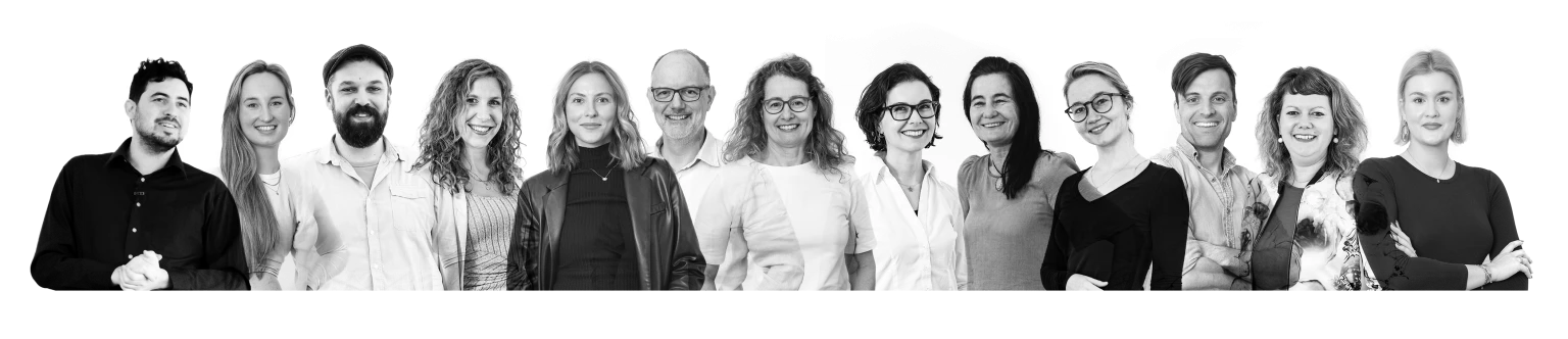 Schwarz-weiß-Gruppenfoto von 14 fröhlichen Menschen unterschiedlichen Alters, die nebeneinander stehen und in die Kamera lächeln.