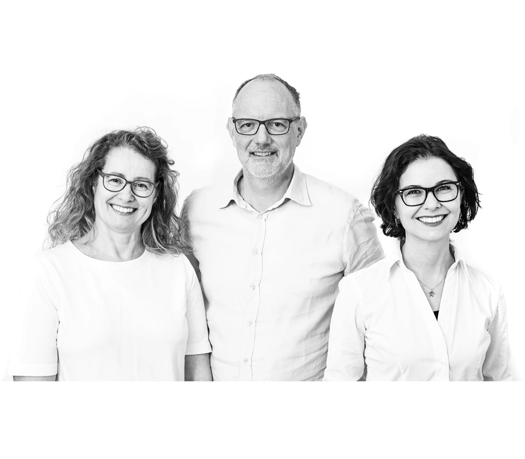 Schwarz-weiß-Foto von drei lächelnden Personen mit Brille. Zwei Frauen stehen links und rechts von einem Mann in der Mitte, alle drei tragen helle Oberteile.