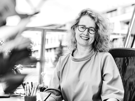 Schwarz-weiß-Porträt einer lächelnden Frau mit lockigen Haaren und Brille, die leicht schräg zur Kamera sitzt. Im Hintergrund sind unscharf Gebäude und Wasser zu erkennen.