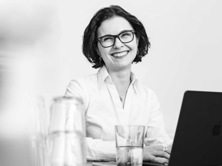 Lächelnde Frau mit Brille und dunklem Haar in weißer Bluse vor hellem Hintergrund.