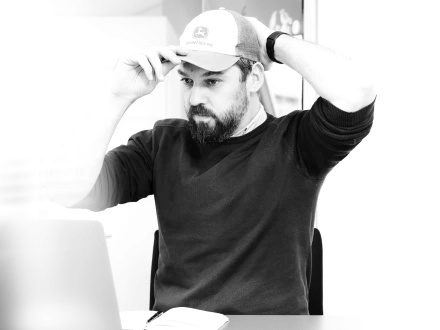 Mann mit Bart und dunklem Pullover setzt sich eine Kappe auf und blickt konzentriert auf einen Laptop.