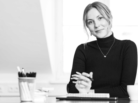 Frau mit zurückgebundenen Haaren und dunklem Rollkragenpullover blickt freundlich in die Kamera und hält einen Stift in der Hand.