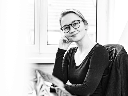 Lächelnde Frau mit Brille und hochgesteckten Haaren stützt den Kopf auf die Hand und sitzt vor einem Fenster.