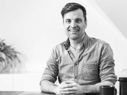 Lächelnder Mann in Jeanshemd blickt in die Kamera und sitzt vor hellem Hintergrund.