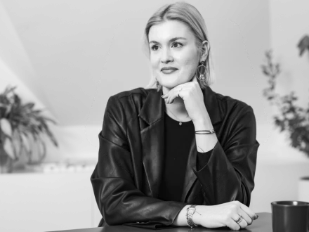 Frau mit kinnlangem, glattem Haar und auffälligen Ohrringen stützt das Kinn auf die Hand und blickt nach rechts.