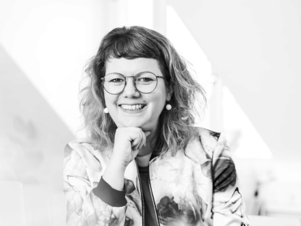 Lächelnde Frau mit lockigem Haar, Brille und auffälligen Ohrringen stützt das Kinn auf die Hand.