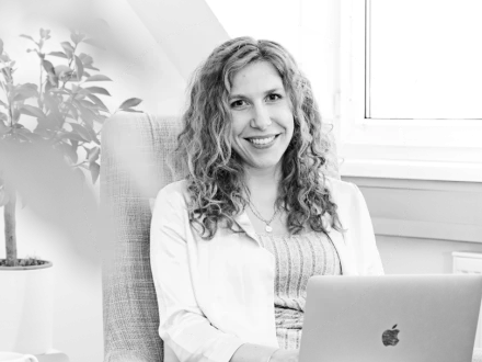 Lächelnde Frau mit lockigem, langen Haar sitzt in einem Sessel vor einem Laptop und blickt in die Kamera.